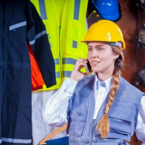 Woman engineer in safety gear with braided hair talks on phone in industrial setting.