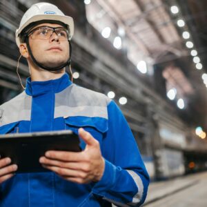 Factory worker in safety gear using a tablet in a well-lit industrial setting.