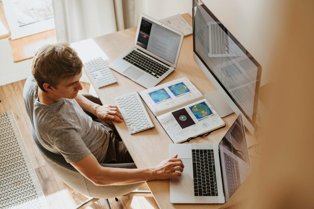 News A man multitasks with laptops and a desktop, coding in a home office setting.
